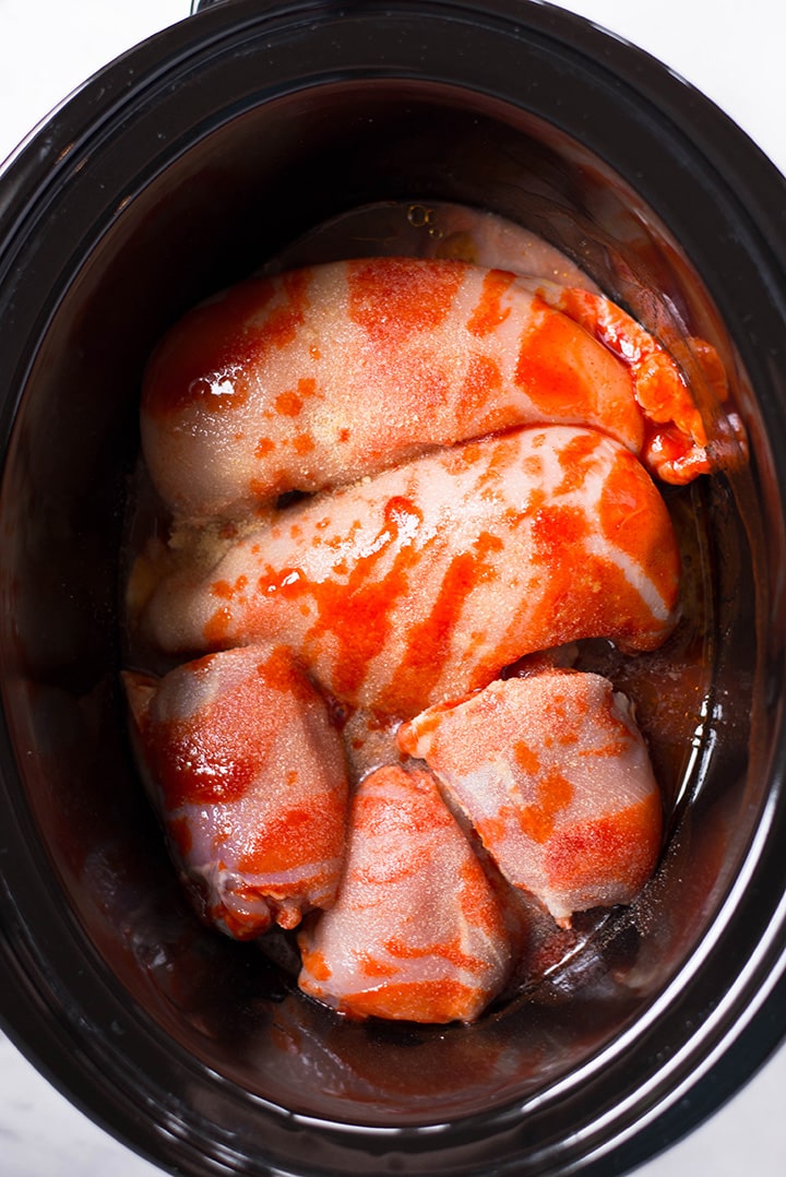 Making Slow Cooker Buffalo Shredded Chicken in the slow cooker. In the slow cooker can be seen chicken breast and chicken thighs in the slow cooker seasoned with hot sauce, olive oil, maple syrup, cider vinegar, garlic powder, onion powder, and sea salt.