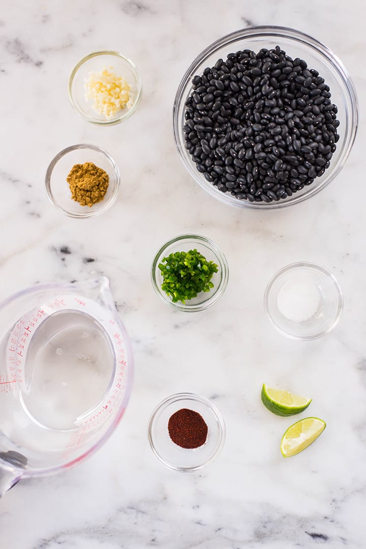 Separated ingredients for refried black beans including black beans, water, olive oil, cumin, chipotle chili powder, garlic cloves, jalapeno, sea salt, lime .