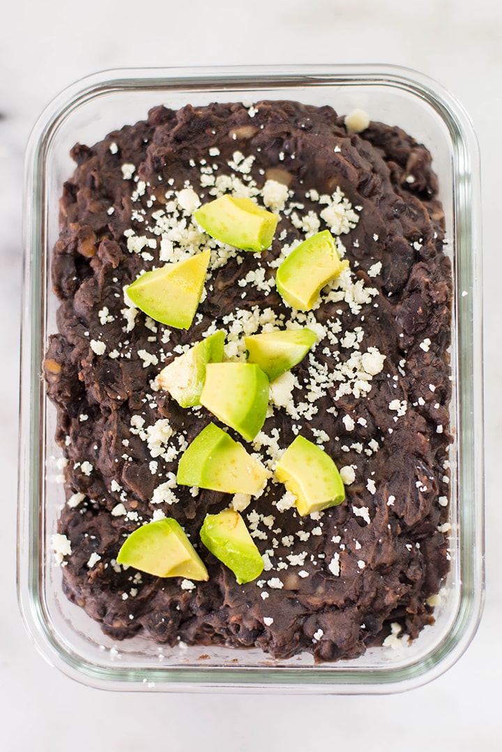 Overhead image of refried spicy black beans topped with avocado in a meal prep container., as a vegan protein dish.