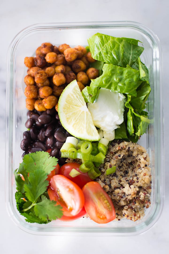 View from the top of a meal prep container filled with the ingredients for the Vegetarian Meal Prep Recipe including baked chickpeas, cooked quinoa, baby greens, grape tomatoes, cilantro, lime slices, and black beans.