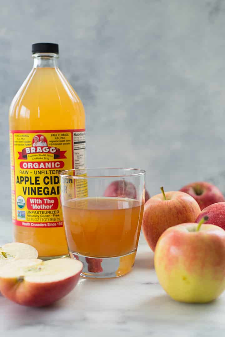 A glass filled with apple cider vinegar next to the bottle of organic apple cider vinegar surrounded by apples.