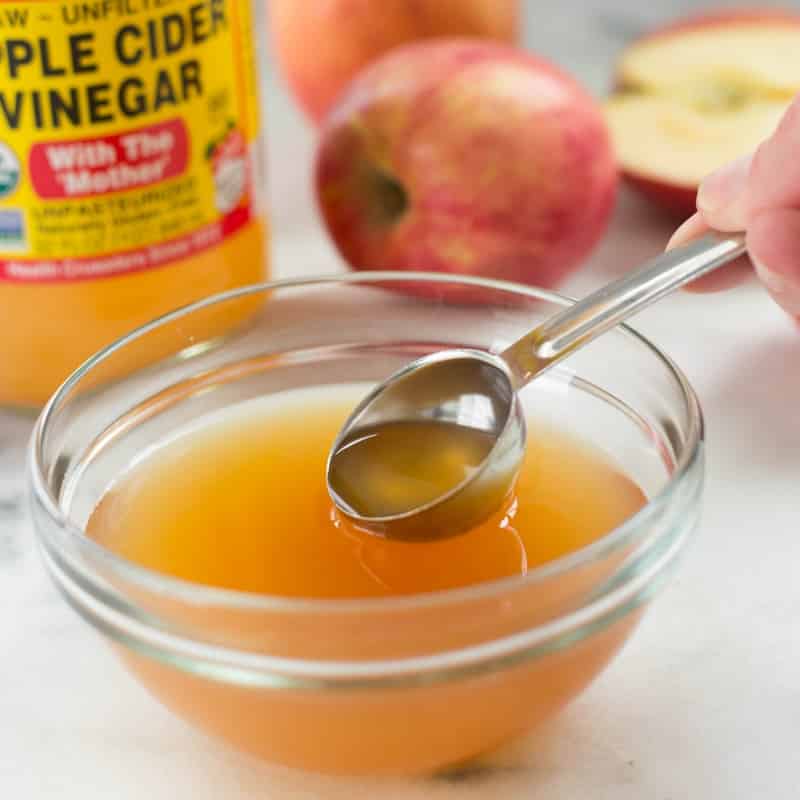 Close up image of a bowl of apple cider vinegar, with a teaspoon being dipped into the bowl. Behind is a bottle of apple cider vinegar and some apples.