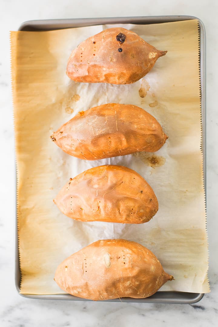Four sweet potatoes on a parchment paper lined baking sheet, baked and ready to be made into The Best Healthy Potato Skins.