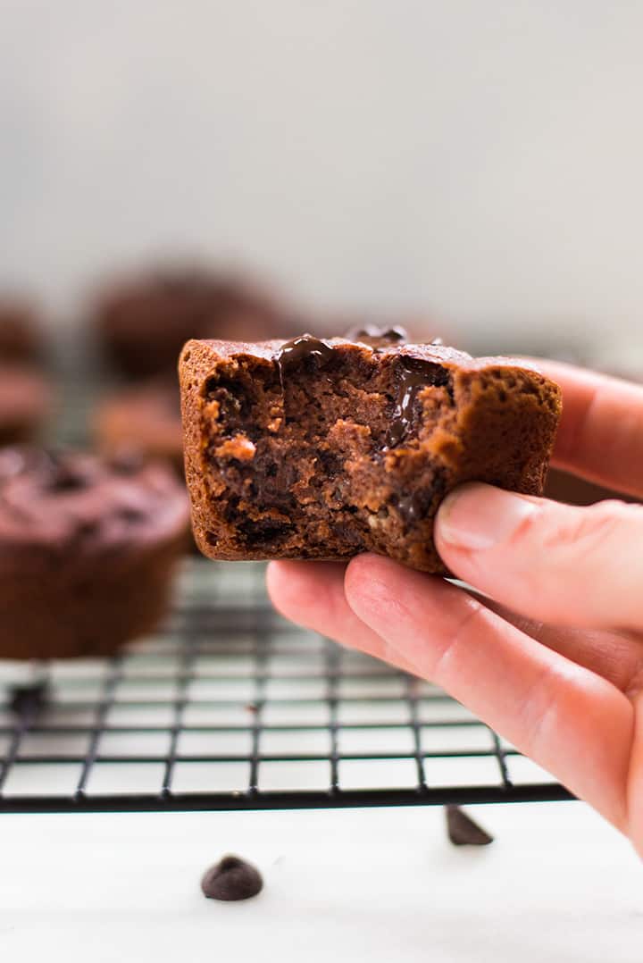 Lacey holding a chocolate muffin with a bite taken out of it. In the background can be seen the cooling rack with other muffins.