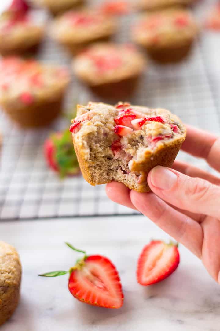 A hand holding a strawberry muffin with a bite taken out of it. In the background can be seen the cooling rack with other strawberry muffins and a few fresh strawberries.