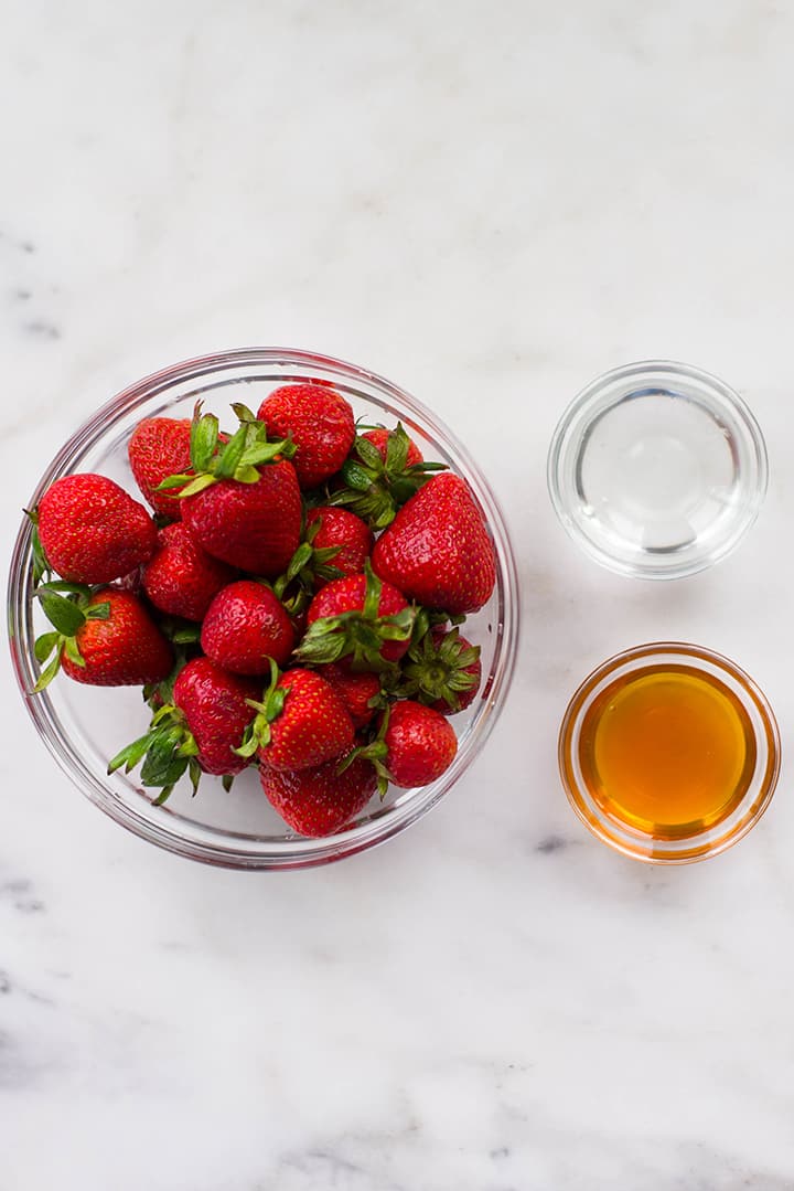 Separated ingredients for strawberry sorbet including fresh strawberries, raw honey, and warm water.