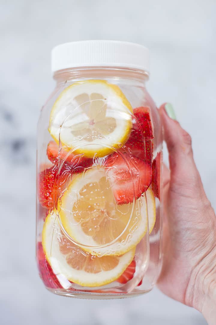 Hand holding a mason jar filled with water, strawberry slices, and lemon slices.