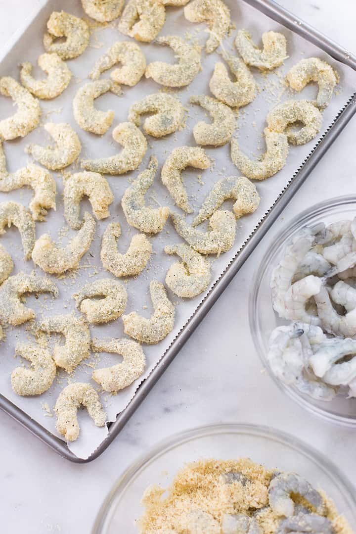 Overhead view of rimmed baking sheet with breaded shrimp, ready to bake. Also in image is more un-breaded shrimp and the almond meal coating.