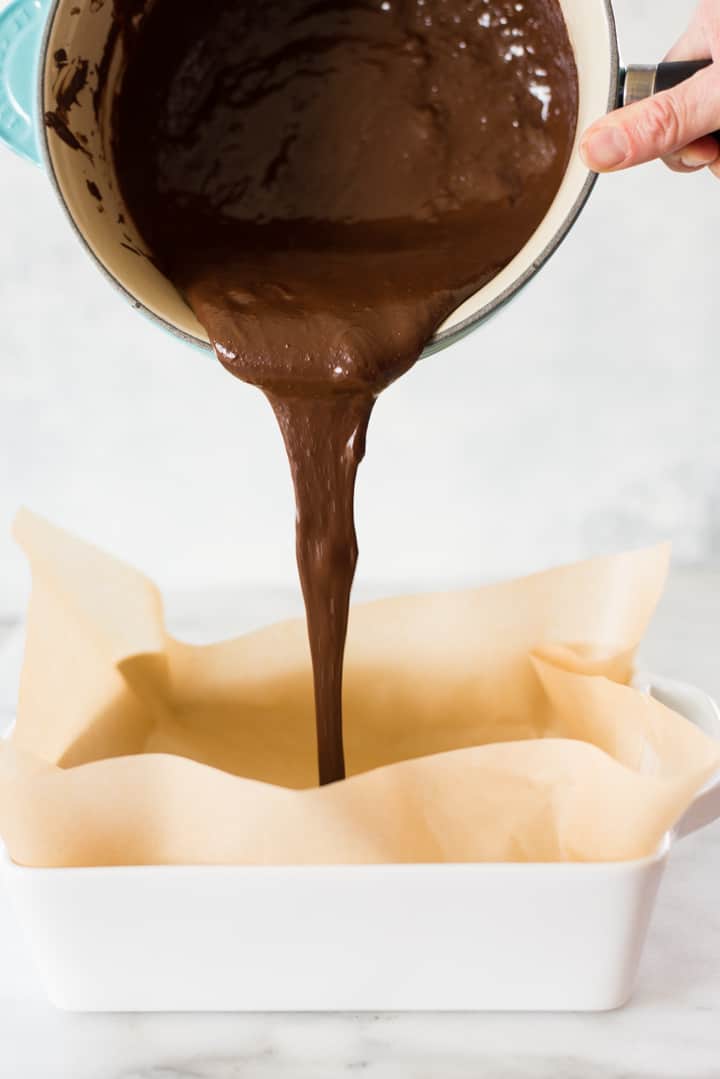 Hand holding saucepan filled with melted dark chocolate mixture that is being poured into an 8x8-inch casserole dish lined with parchment paper to create the bottom layer of the Easy Chocolate Peanut Butter Fudge.
