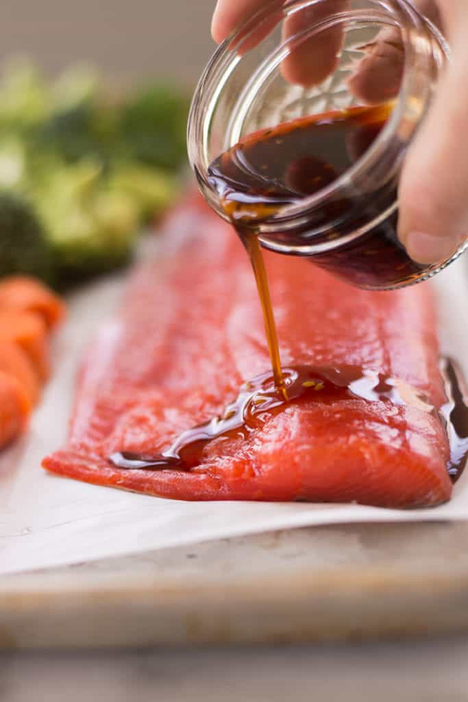 Side view of a small jar of the best healthy teriyaki sauce bring poured over a piece of salmon, in preparation for cooking.
