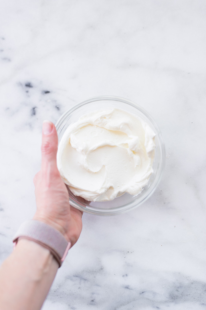 Overhead image of a hand holding a clear glass bowl of plain Greek yogurt, a healthy alternative to oil in baking.