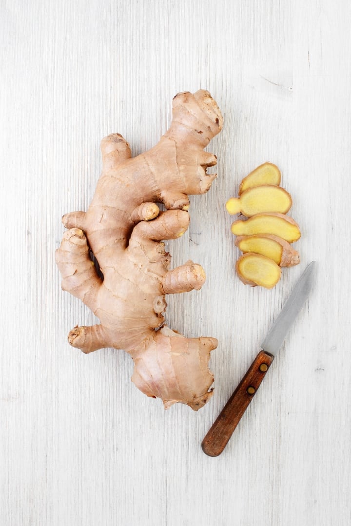 Close up overhead view of ginger on a cutting board, with a knife and cut ginger slices off to the side.