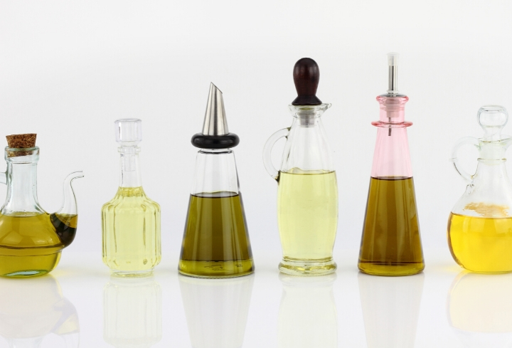 Side view of 6 different shaped bottles, filled with various types of cooking oil, all lined in a row.