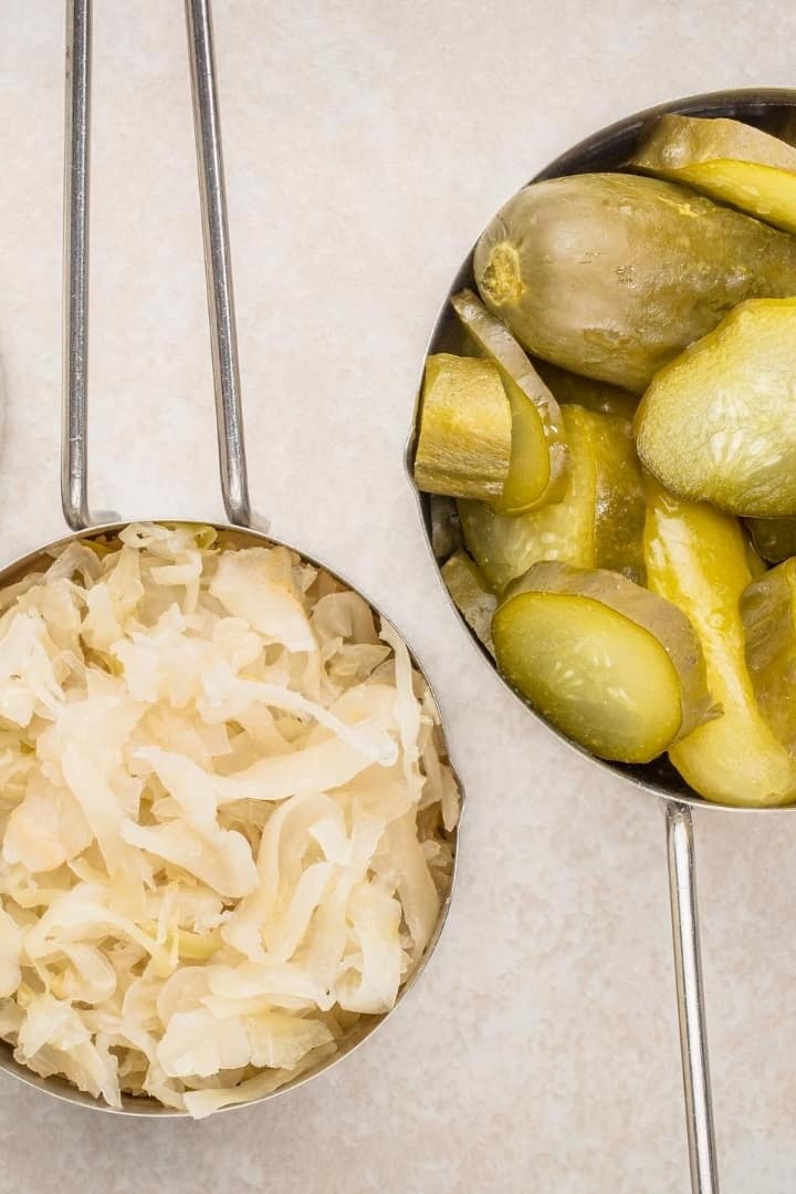 Overhead view of two small metal serving dishes, one filled with sauerkraut and one with pickles, both fermented foods.