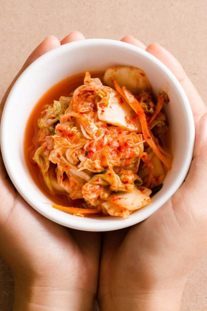 Close up view of two hands holding a white bowl filled with kimchi.