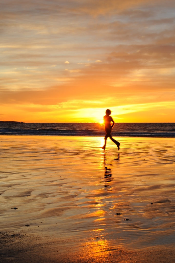 Image of a person running along a beach with the sun setting behind them.