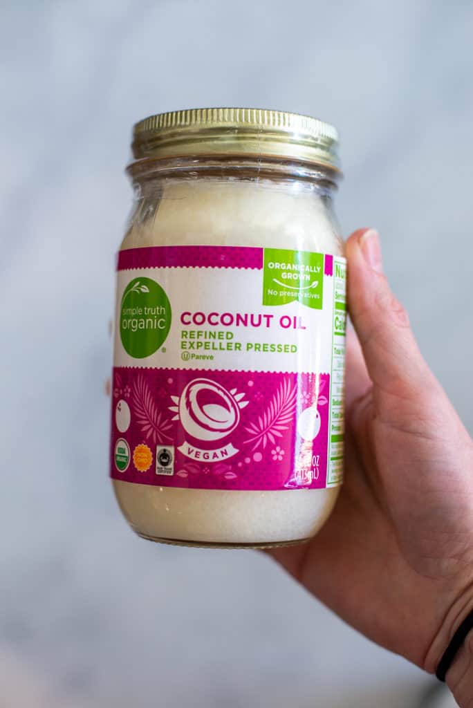 Close up side view of a hand holding a large jar of coconut oil with a pink label.