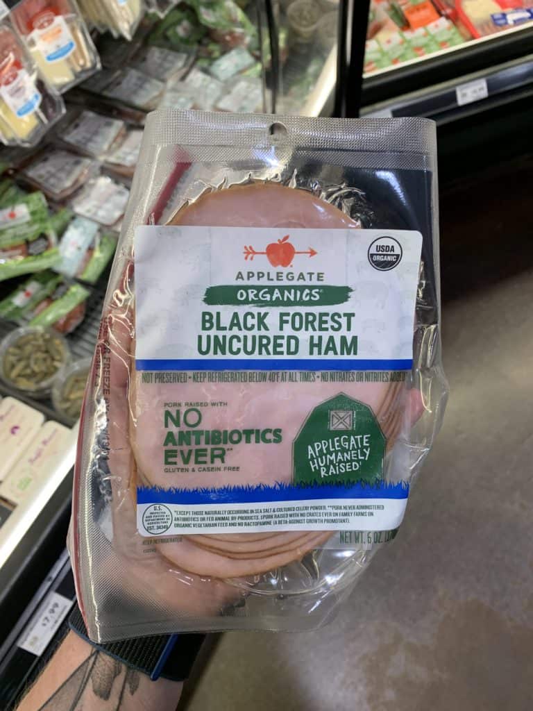 Close up image of a hand holding a package of deli meat, uncured black forest ham.