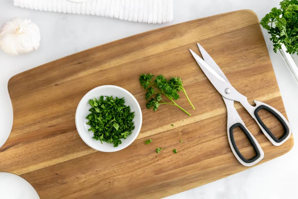 Overhead image of a cutting board with scissors and Italian parsley on it, with some parsley in a white bowl.