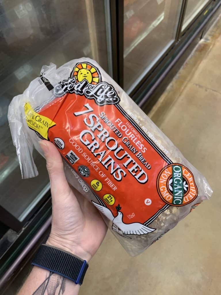 Close up image of a hand holding a loaf of Food for Life Bread with a clear and orange label.
