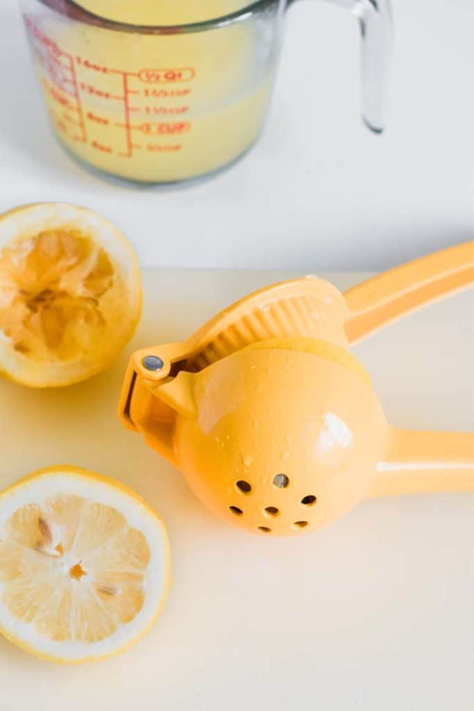 Close up view of a lemon juicer, with a measuring cup with lemon juice in it, a squeezed lemon, and half an unsqueezed lemon beside it.