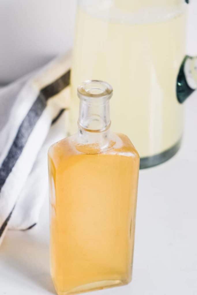 Close up image of a bottle of simple syrup, some of which will be poured into the lemonade pitcher behind it.
