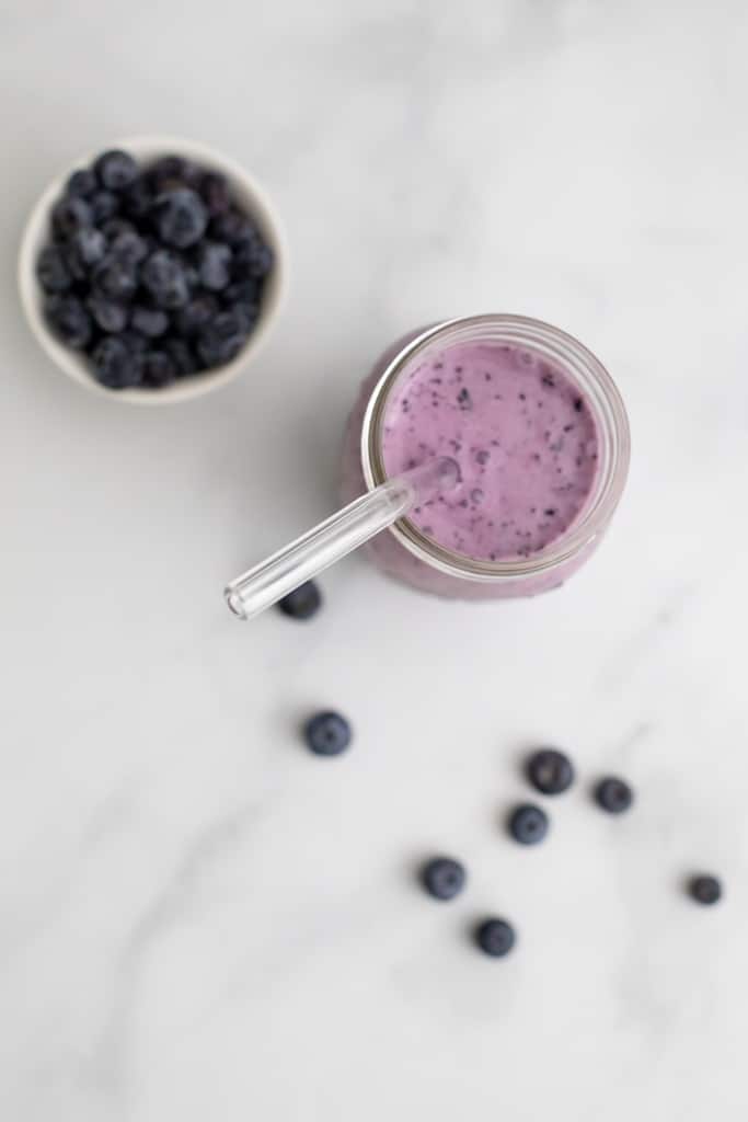 Close up overhead view of a mason jar filled with the Blueberry Protein Smoothie, with blueberries in a bowl beside it.
