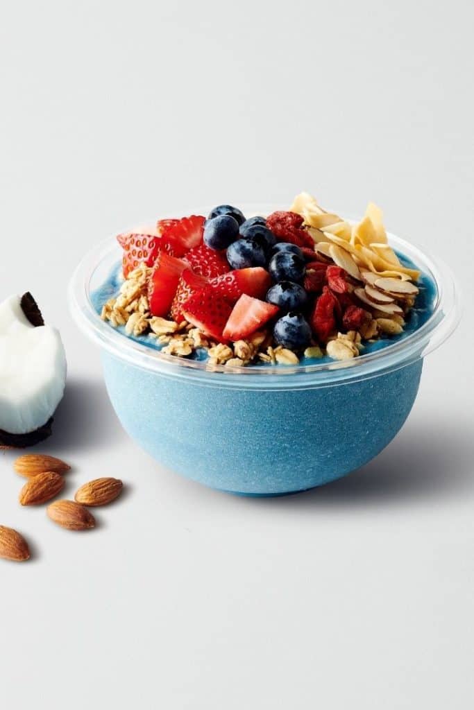 Close up side image of a blue bowl with granola, strawberries, blueberries, and almond slices, with almonds and coconut beside it.