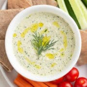 A close-up of a bowl of tzatziki sauce with a drizzle of olive oil, garnished with fresh dill, and paired with pita bread and raw vegetables for dipping.