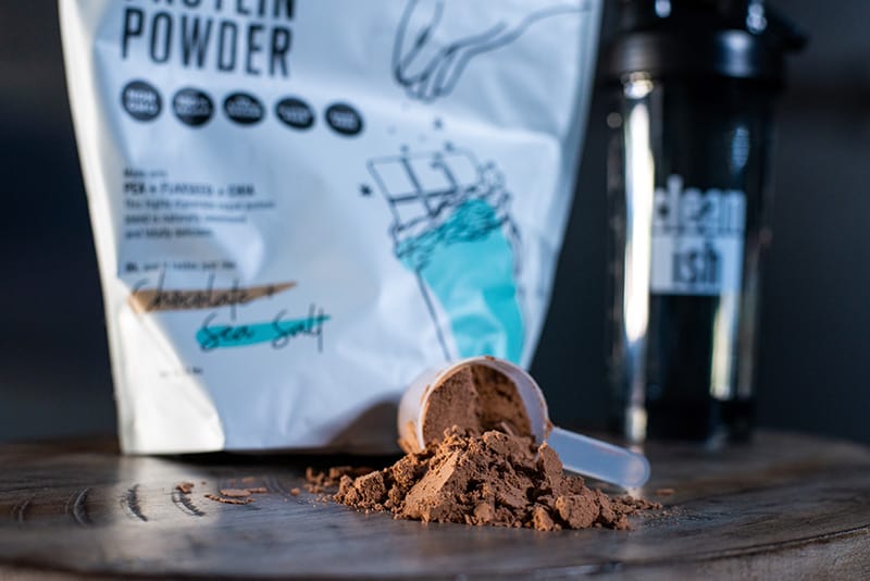 Close up image of Chocolate and Sea Salt Protein Powder, with a scoop of powder beside it and a Cleanish Bottle behind it.