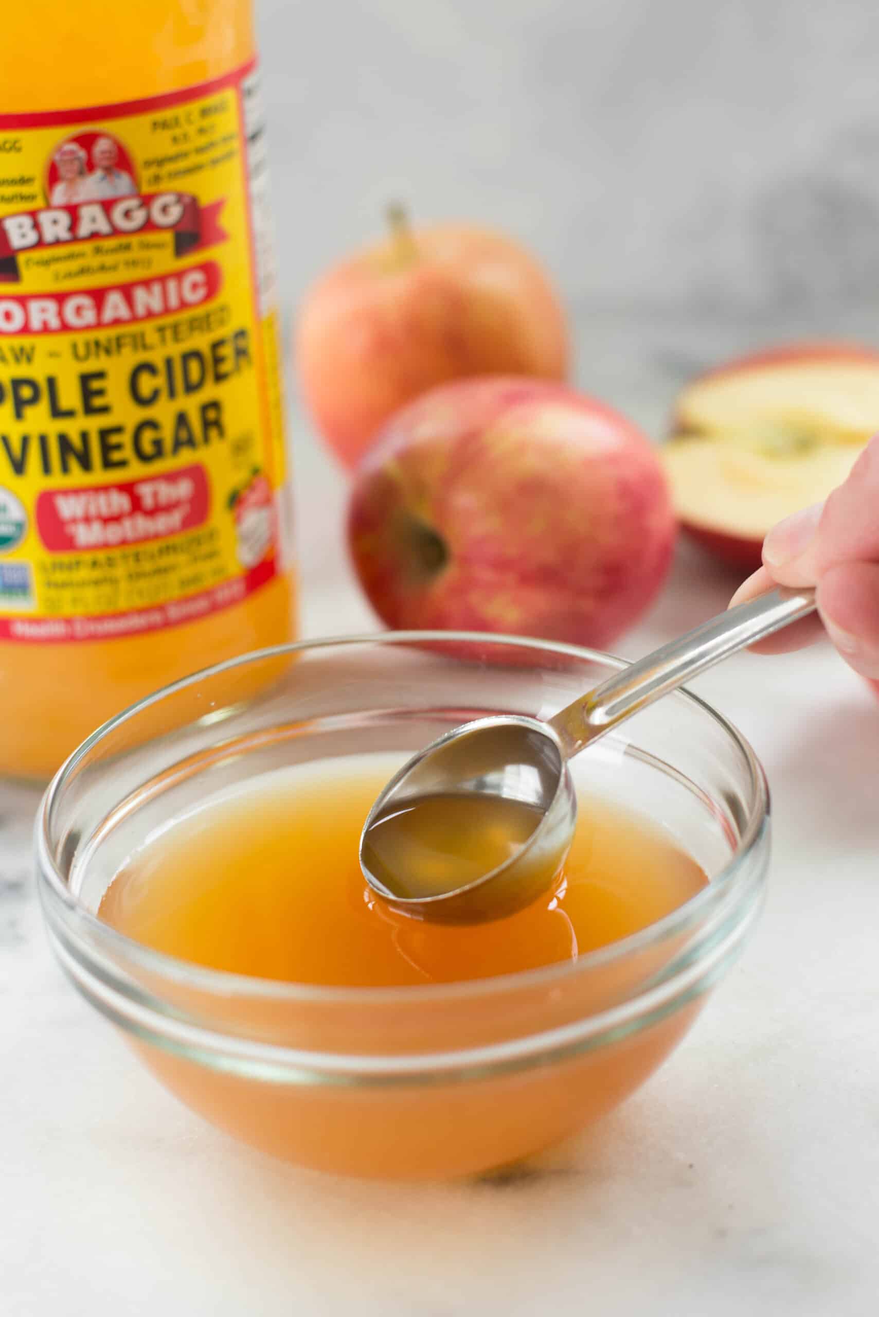 Taking a tablespoon of apple cider vinegar from a bowl filled with apple cider vinegar. In the background can be seen the apple cider vinegar bottle and some apples.