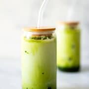 A 1200x1200 photo of a iced matcha collagen latte in front of a white marble background with a blurred matcha latte in the back.
