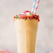 Vanilla cupcake protein smoothie in a tall glass topped with whipped cream and rainbow sprinkles, with striped paper straws